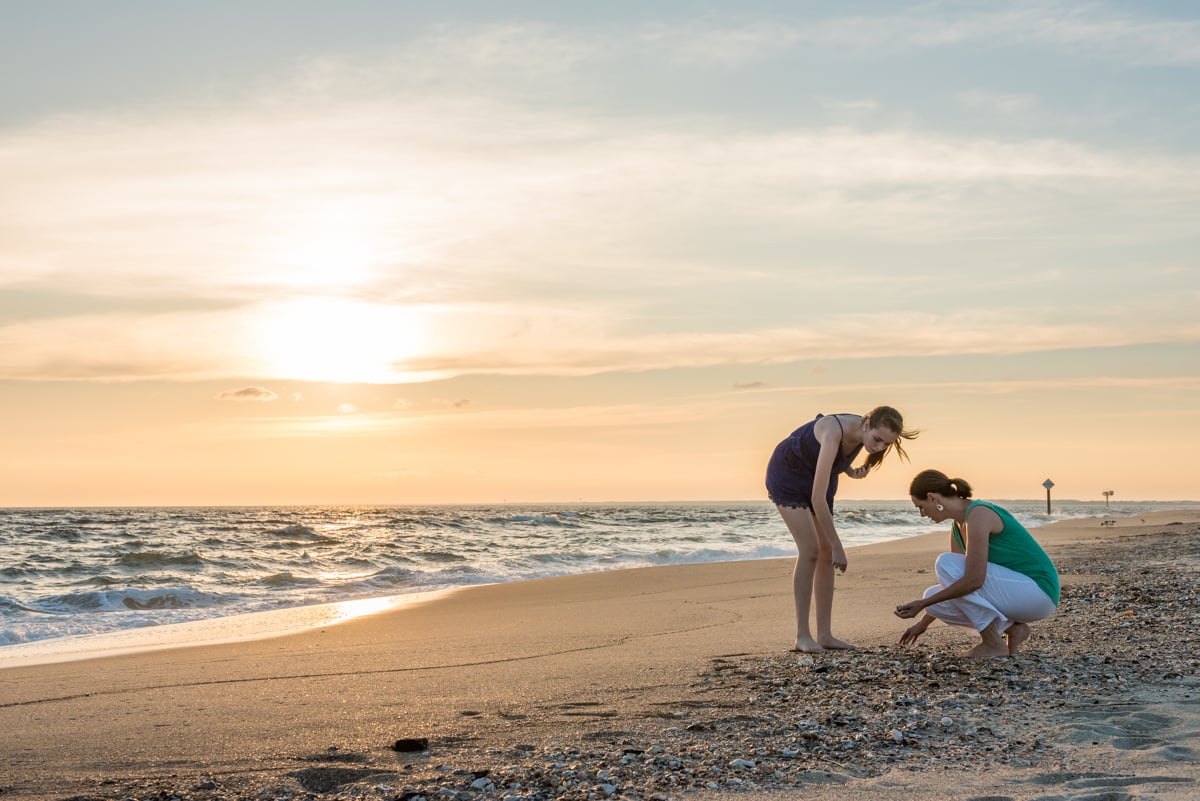 An image of two woman collecting seashells on a beach at sunset.